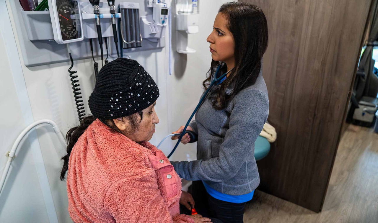 A female doctor takes a blood pressure reading of an older patient inside a mobile health van with medical equipment.