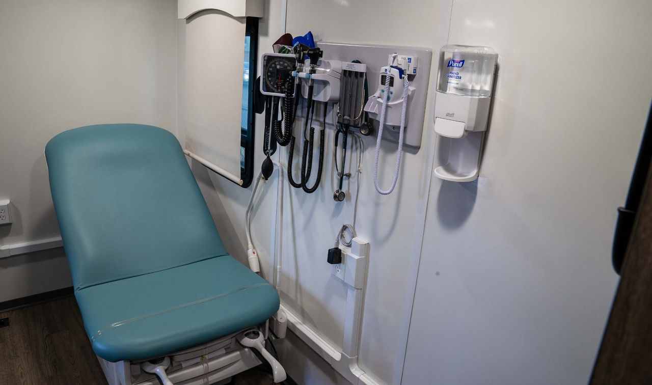 A medical examination area in a mobile van with medical equipment and sanitizing gel on the wall.