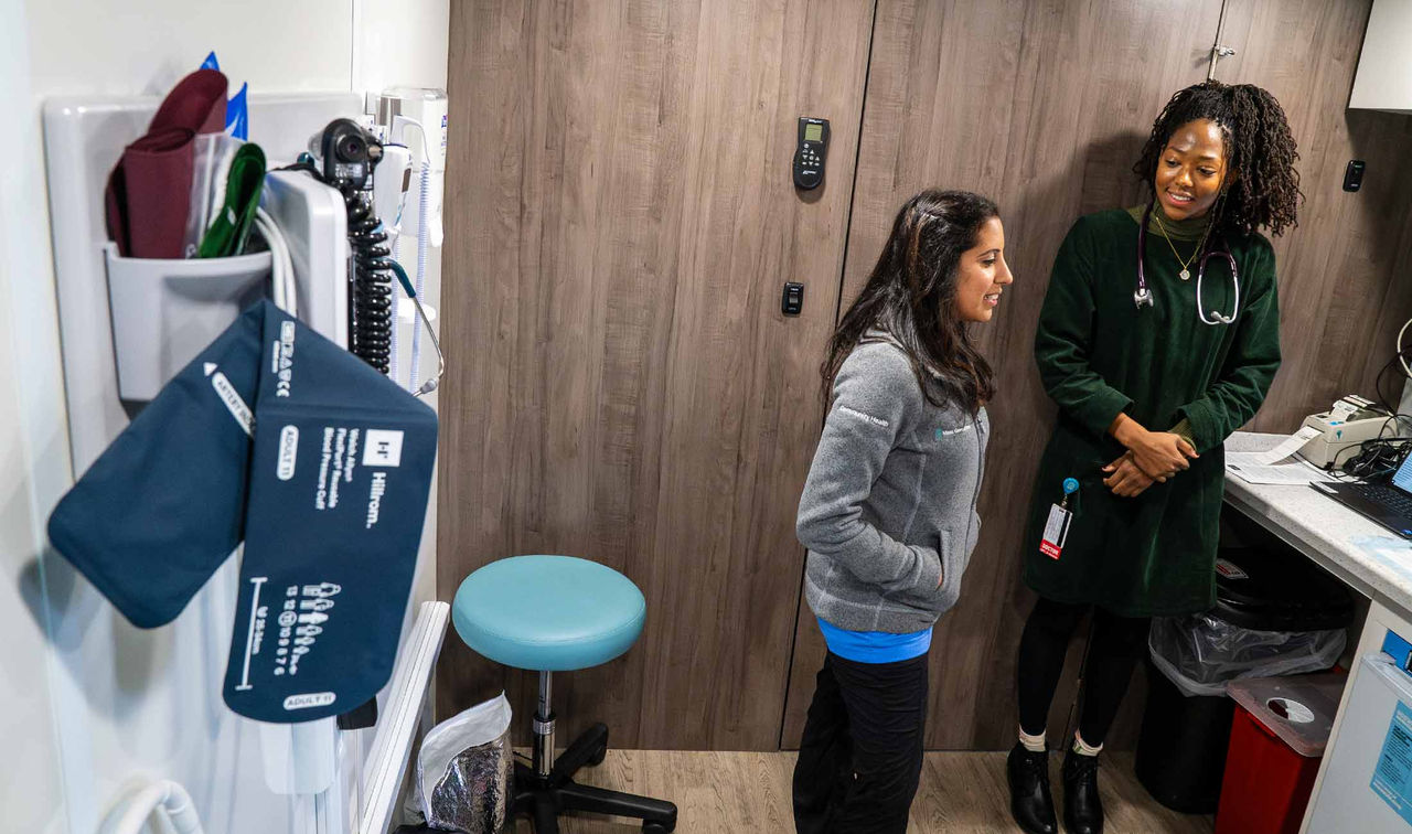 A blood pressure cuff inside a mobile health van with two female doctors standing, smiling, and chatting in the background