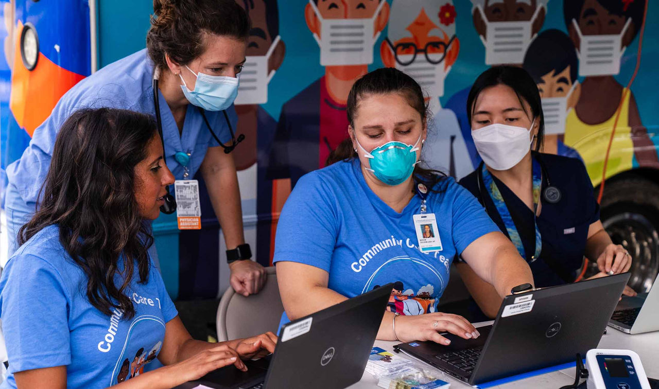 Four healthcare professionals sitting and standing outside the Mass General Brigham Community Care Van, looking at two laptop screens and having a discussion. Two of them are wearing blue scrubs with lanyard tags, while the other two are wearing blue short-sleeved t-shirts that say "Community Care Vans" on the front. Three of them are masked.