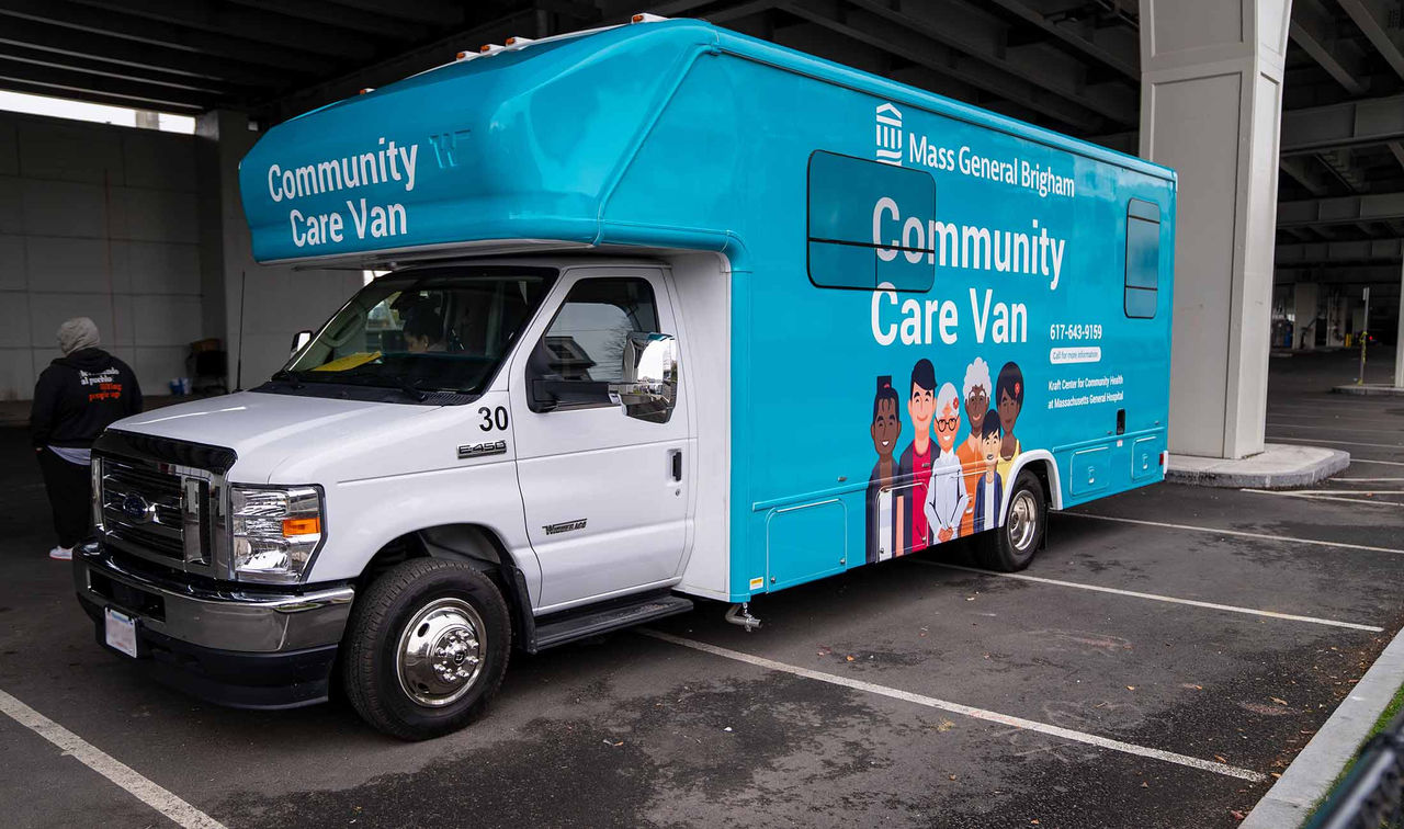 A full front and side view of the Mass General Brigham Community Care Van parked at a location. The van is wrapped in teal on the side consistent with the Mass General Brigham brand colors. The side panel has the Mass General Brigham logo on top with the words "Community Care Van" in large font and phone numbers in smaller font. Part of the side wrap design includes 5 illustrated individuals of varying genders, ethnicities, and ages.