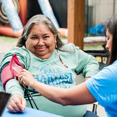 A woman with gray hair while wearing a long-sleeved light green t-shirt. She has a blood pressure cuff on her left arm as a healthcare professional takes a blood pressure reading.