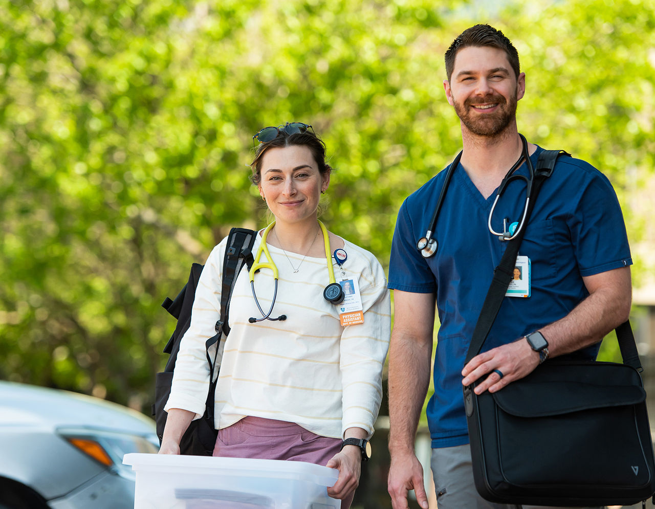 Two providers in an outdoor setting, smiling at the camera. They are wearing scrubs and carrying medical equipment.