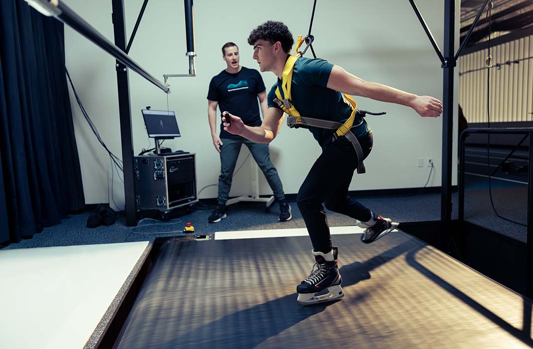 A young male athlete wearing ice skates is training on a treadmill-like platform while secured with a harness. 