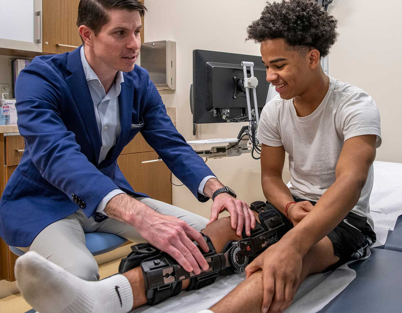 An orthopedics provider is seen adjusting a knee brace on a young male patient in a clinical setting.