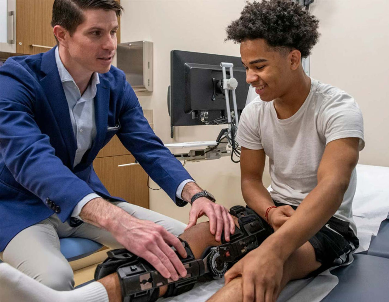 An orthopedics provider is seen adjusting a knee brace on a young male patient in a clinical setting.
