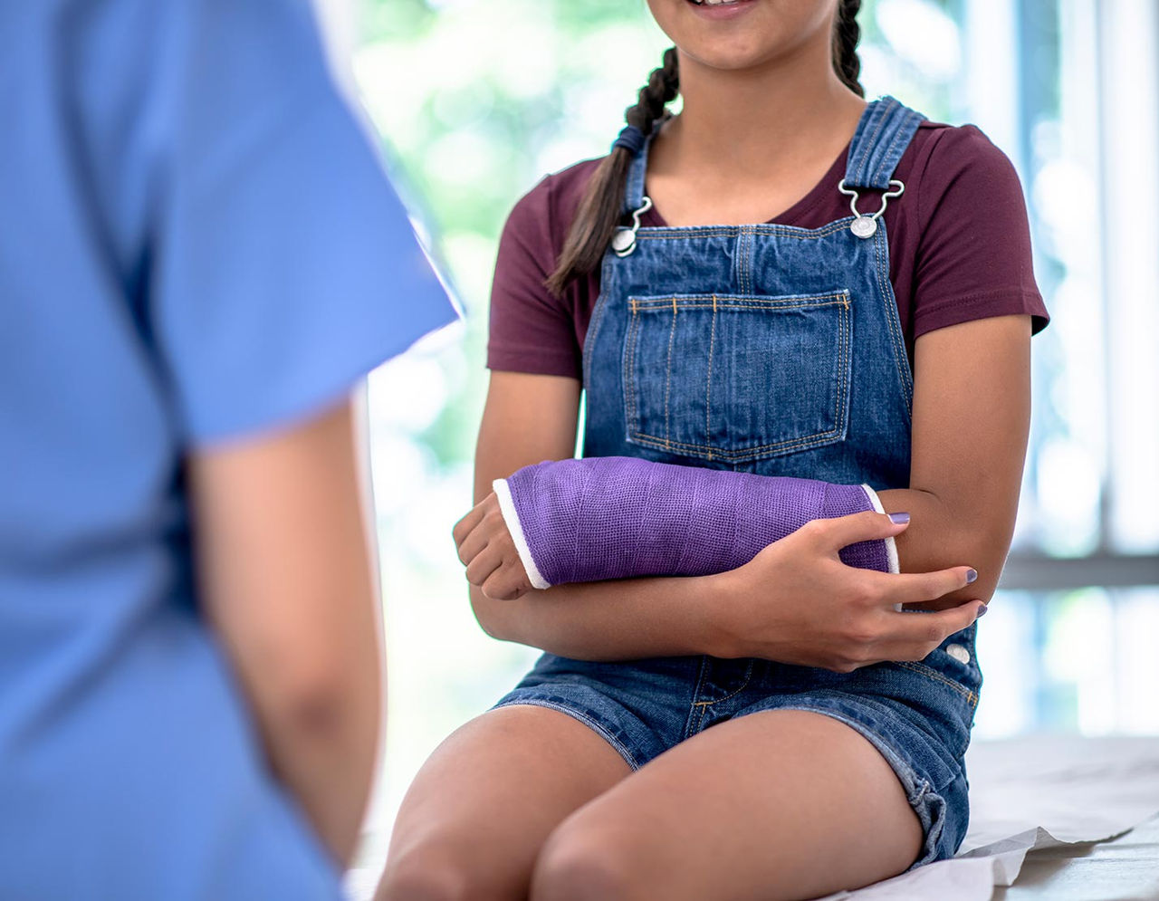 A young girl wearing denim overalls and a maroon shirt sits in a medical clinic with a purple cast on her arm.