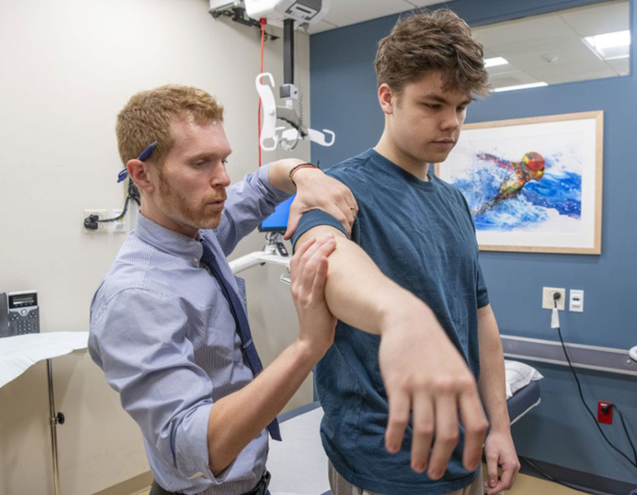 A male clinician assists a patient with arm exercises in exam room.