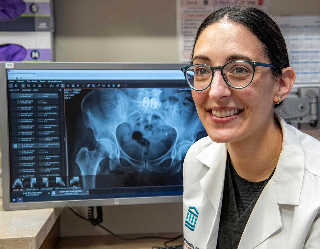 A medical professional is seen in a clinical setting with a computer screen displaying a pelvic X-ray.