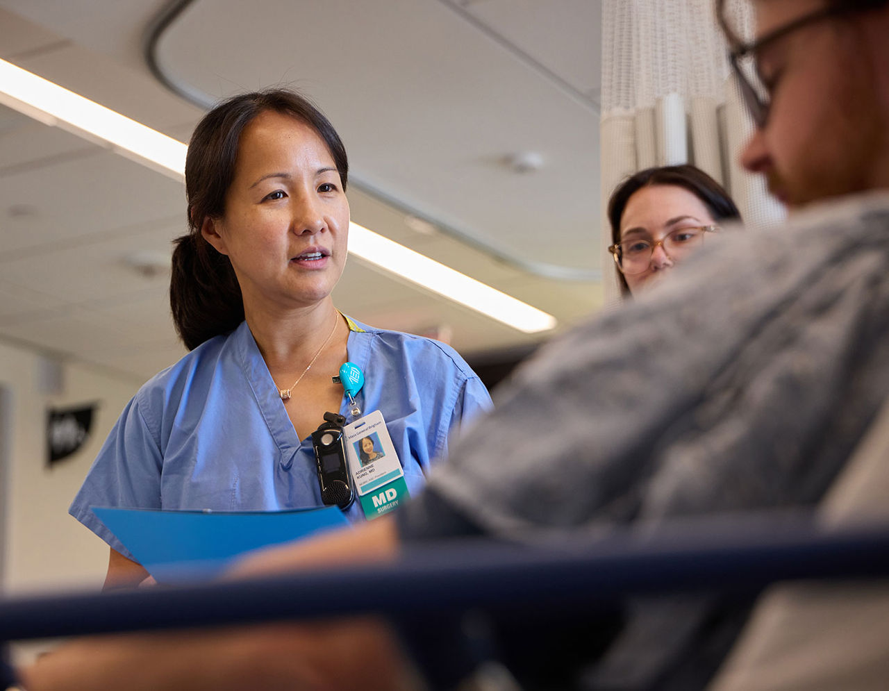 A healthcare worker wearing blue scrubs and an MD badge is seen in a hospital environment next to patient in bed.