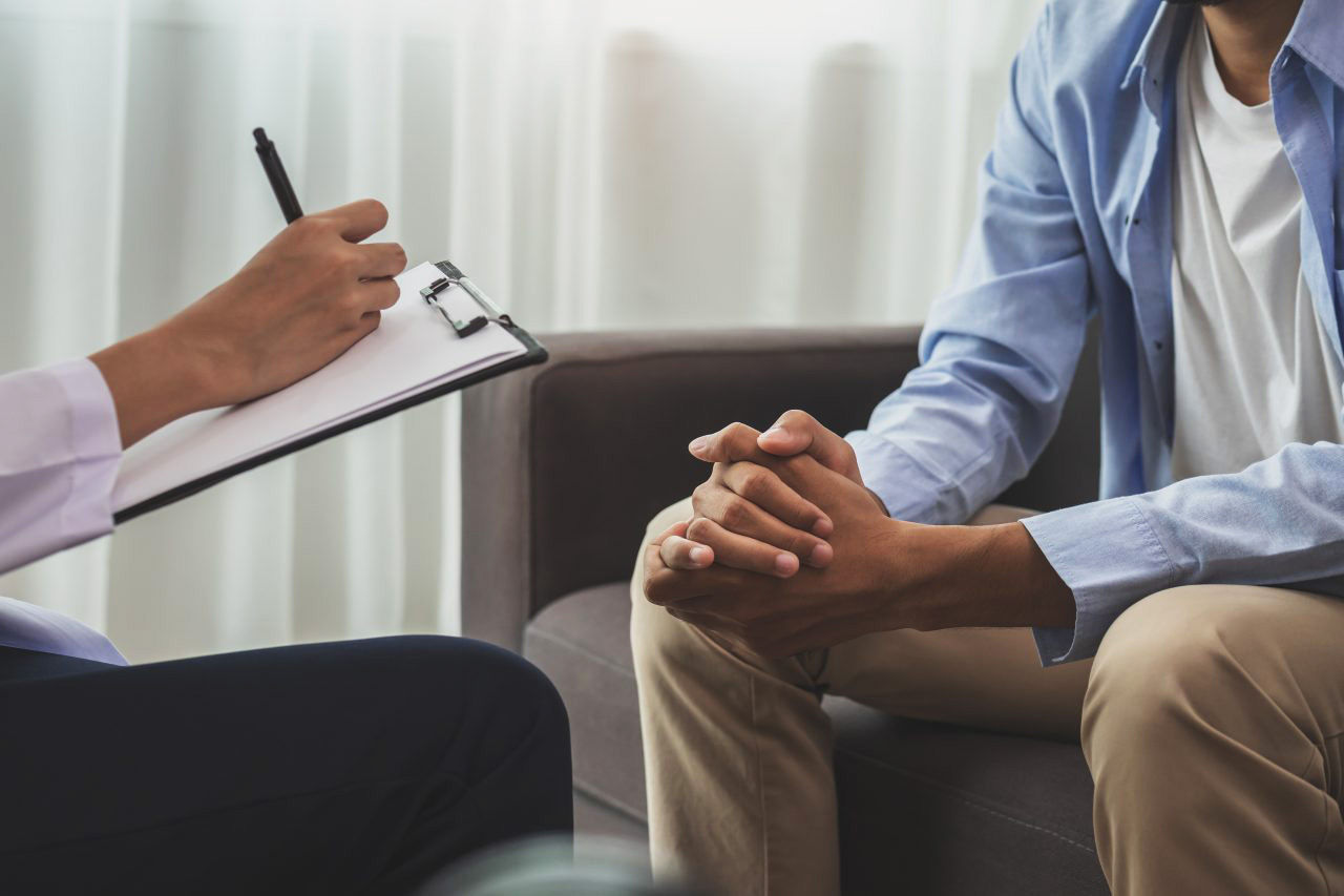 A man sits on a couch with folded hands, talking to a doctor. with a clipboard.