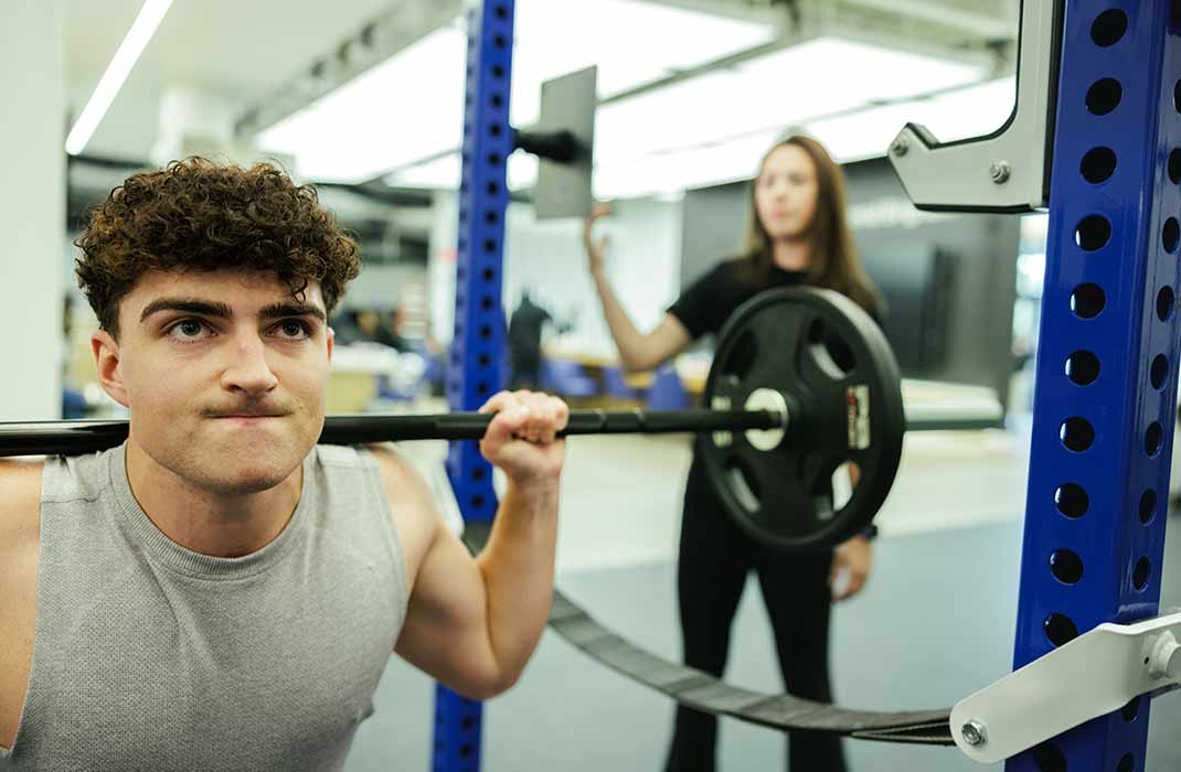 man squatting with barbell on his shoulders with female athletic trainer supervising
