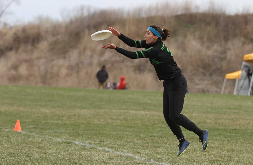 A person is captured mid-air while catching a frisbee on a grassy field. The setting appears to be an outdoor sports event with cones marking the play area. The individual is dressed in athletic attire, and the background includes a hill and a yellow tent. The scene conveys action and focus.