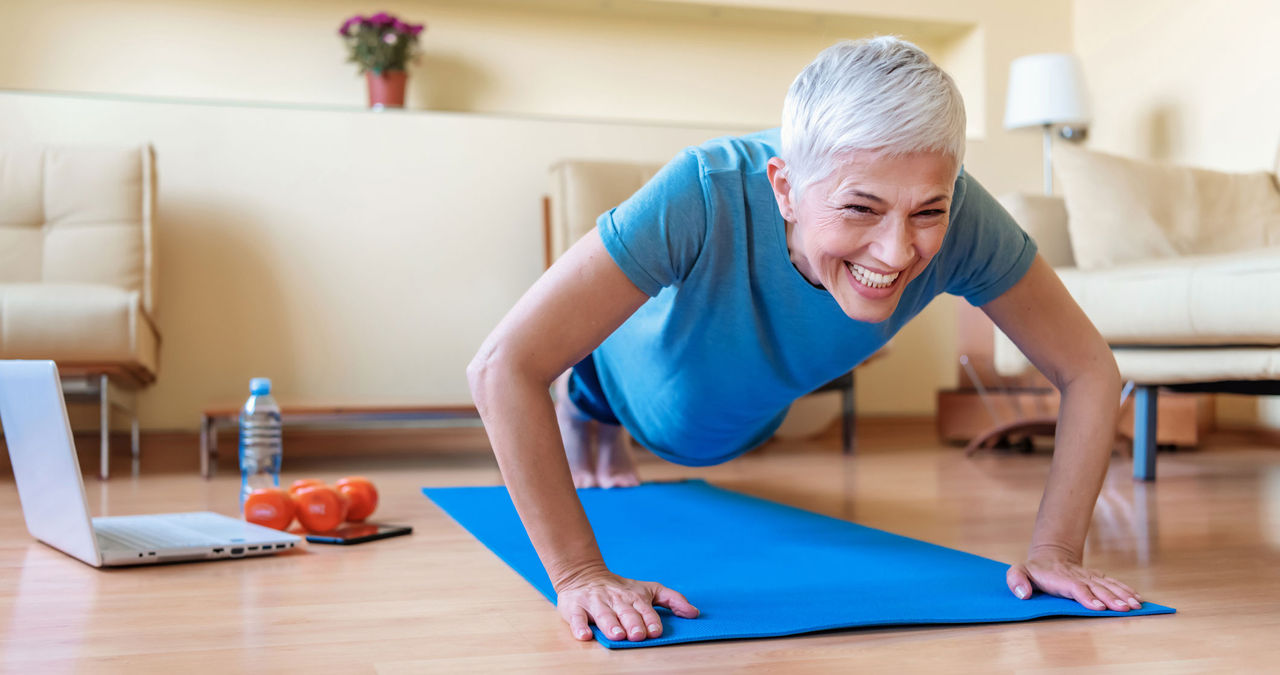 A grinning older woman planks on a yoga mat on her living room floor.