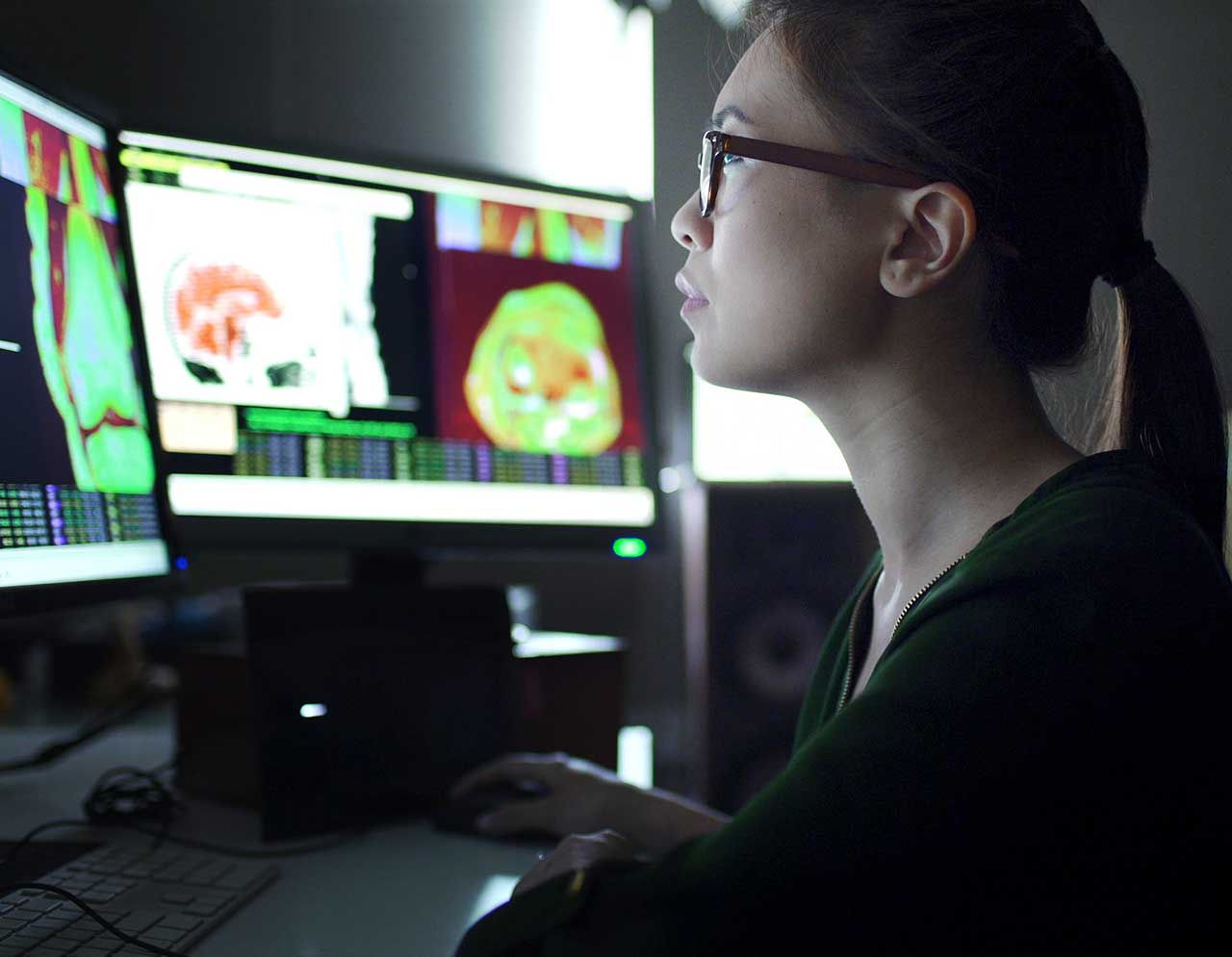 A woman in a darkened room analyzes medical scans on a bank of computer screens.