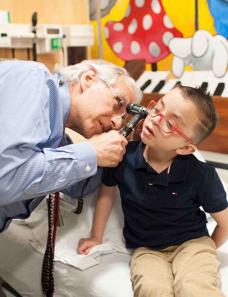An older male doctor uses an otoscope to examine a young boy’s ear in a medical clinic.