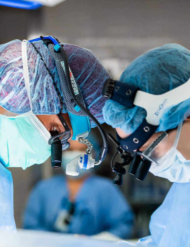 Two surgeons are intensely focused during a surgical procedure, wearing blue scrubs, loupes, masks, and protective headgear.