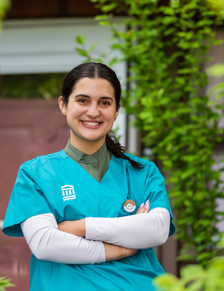 A healthcare professional wearing teal medical scrubs stands with arms crossed in an outdoor setting.