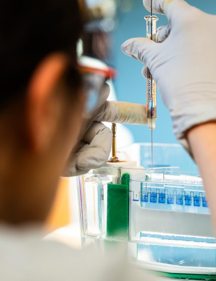 A gloved individual is seen handling a syringe above a laboratory gel electrophoresis apparatus.