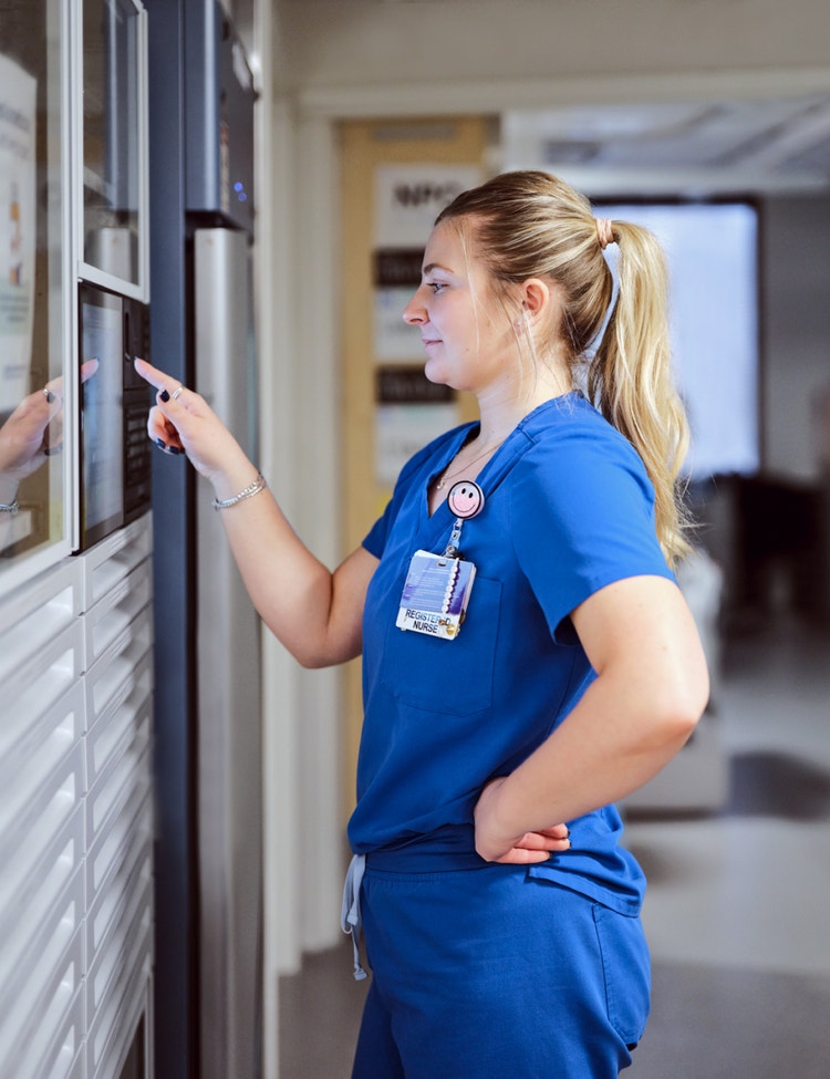 A healthcare professional in blue scrubs interacts with a wall-mounted touchscreen or control panel.