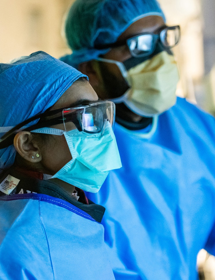 Two medical providers wearing blue surgical gowns, blue hair covers, face masks, and protective eyewear appear to be focused on a task in a surgical setting.
