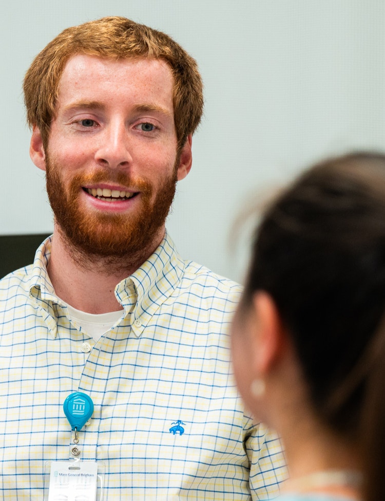 A man wearing a checkered shirt and a blue Mass general Brigham ID badge is engaged in conversation with a woman in an indoor office setting.